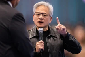 Man in a black leather jacket speaking into a microphone, gesturing with one hand, in a blurred background setting.