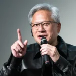 Person in a black leather jacket speaking into a microphone, gesturing with one finger raised.