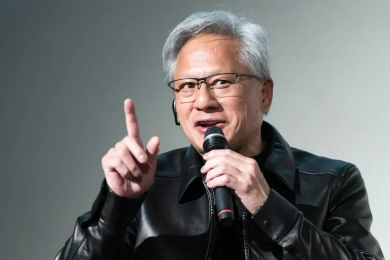 Person in a black leather jacket speaking into a microphone, gesturing with one finger raised.