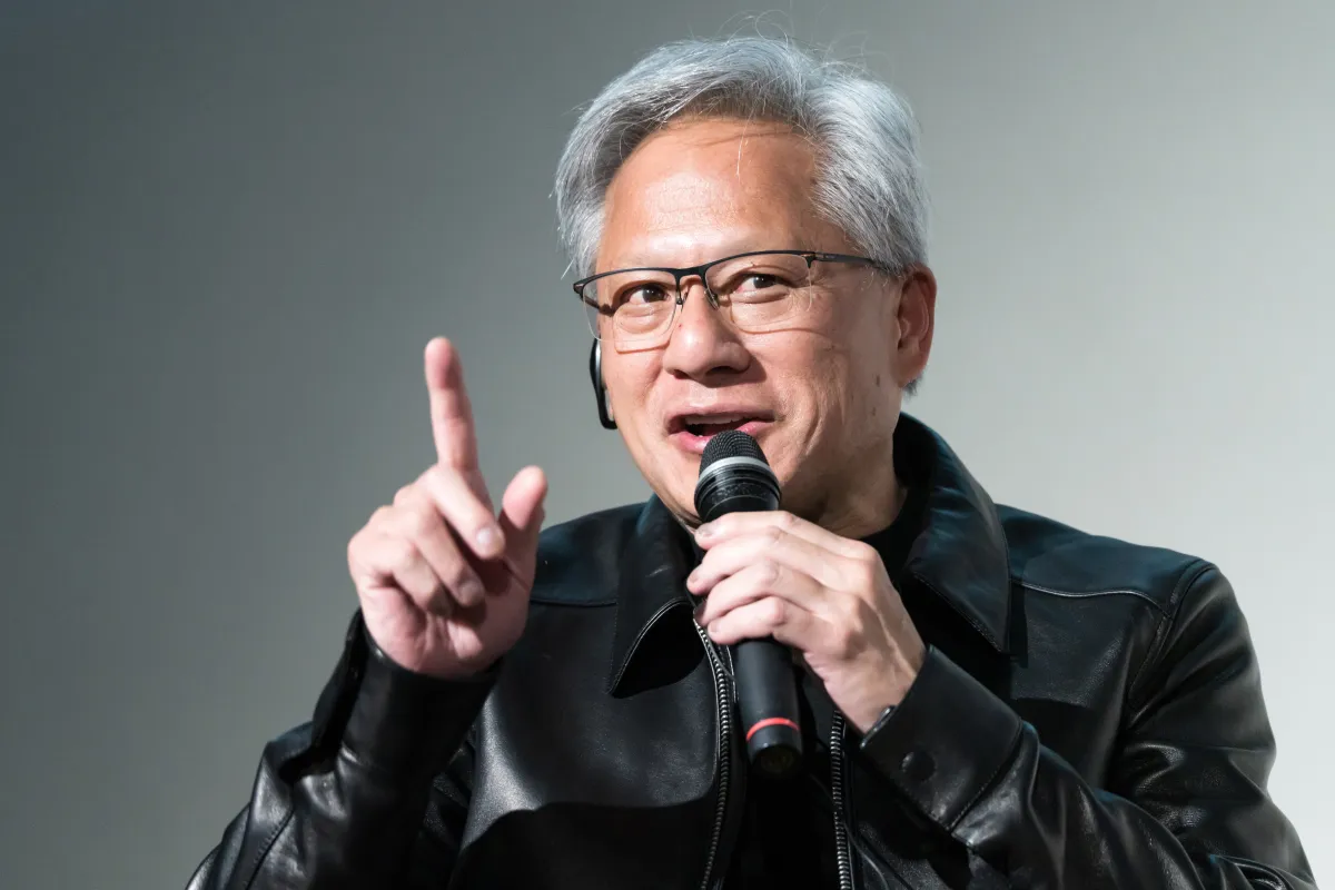 Person in a black leather jacket speaking into a microphone, gesturing with one finger raised.