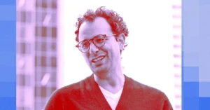 Man with glasses and curly hair in a red-tinted image, standing outdoors with a blurred cityscape in the background.