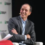 Man in a suit with a headset smiles while sitting on stage, TechCrunch Disrupt logo in the background.