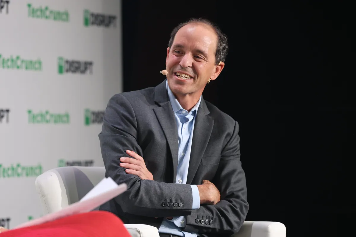 Man in a suit with a headset smiles while sitting on stage, TechCrunch Disrupt logo in the background.