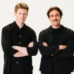 Two men in black shirts stand with arms crossed against a plain white background.