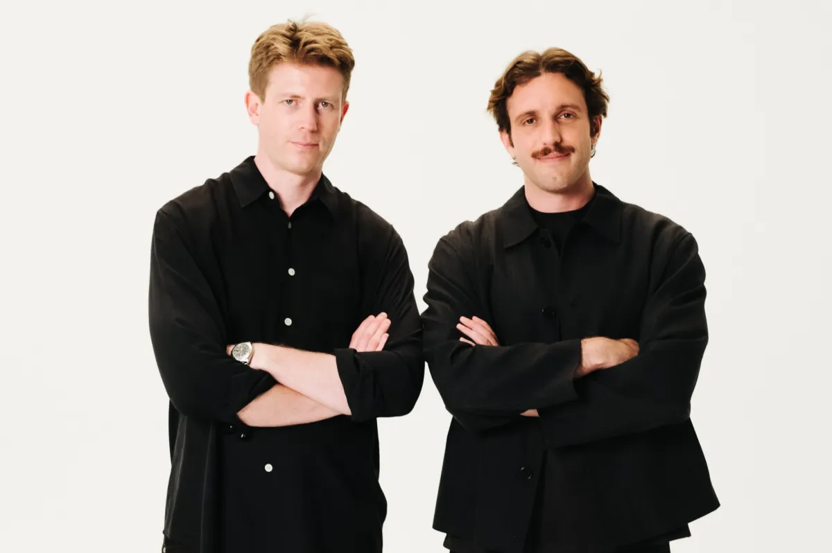 Two men in black shirts stand with arms crossed against a plain white background.