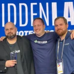 Three men smiling, standing together in front of a HiddenLayer backdrop, one holding an award, all wearing HiddenLayer shi...