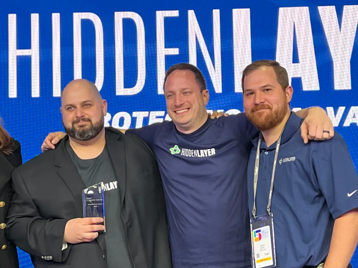 Three men smiling, standing together in front of a HiddenLayer backdrop, one holding an award, all wearing HiddenLayer shi...