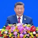 Man in a blue suit speaks at a podium adorned with colorful flowers against a blue background.