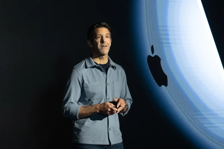 Person speaking on stage with Apple logo in the background, wearing a blue shirt, hands clasped.