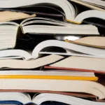 Stack of open and closed books with visible spines and pages, some books overlapping, a yellow pencil among them.