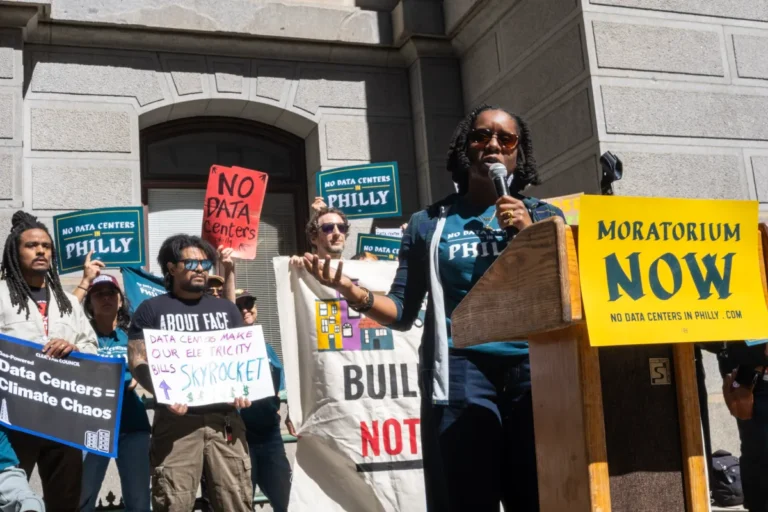 Protesters at City Hall holding signs against data centers, speaker at podium with "Moratorium Now" sign.