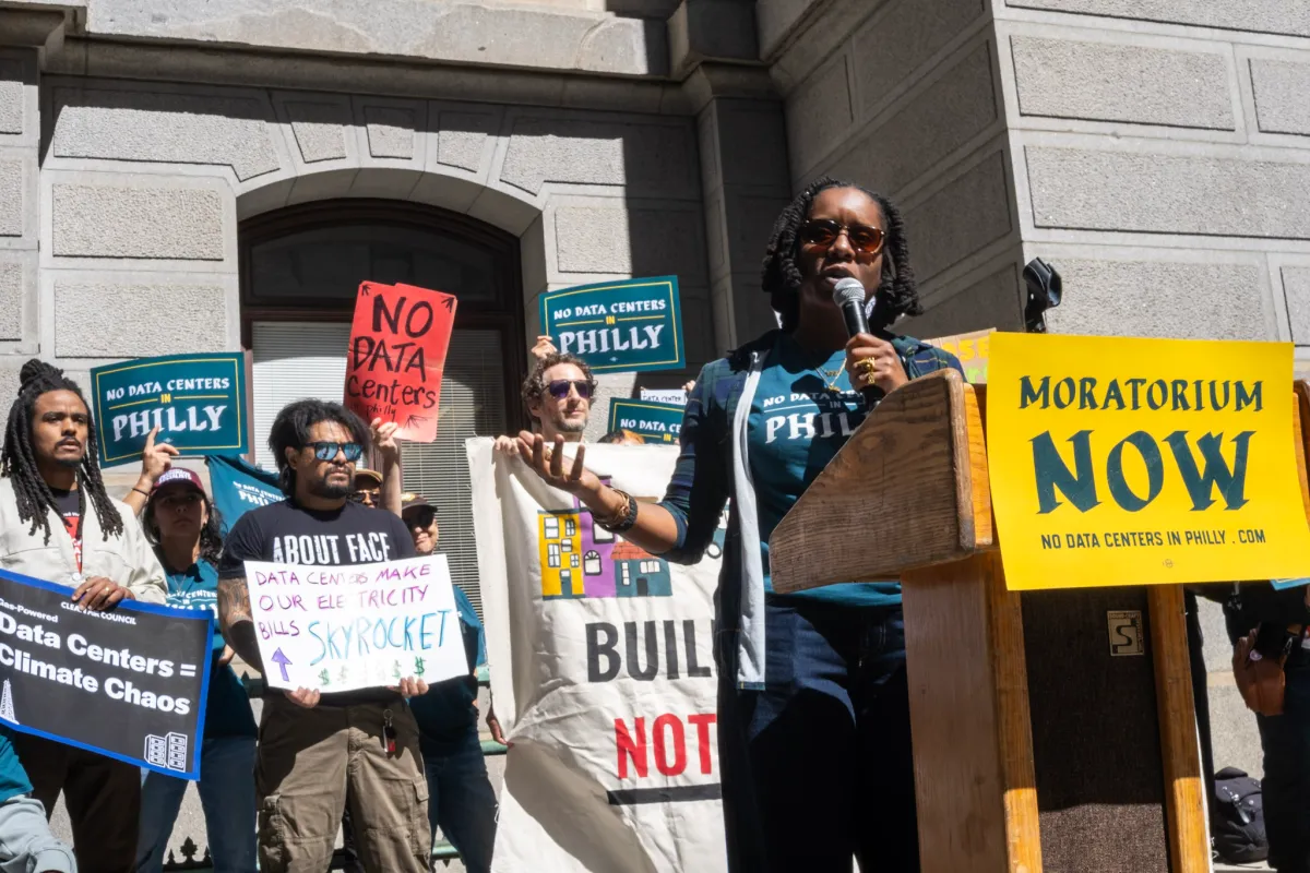 Protesters at City Hall holding signs against data centers, speaker at podium with "Moratorium Now" sign.