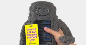 Gray plush toy resembling a server with a smiling face, held by a hand, featuring a yellow tag reading "Hug me for ear pie...