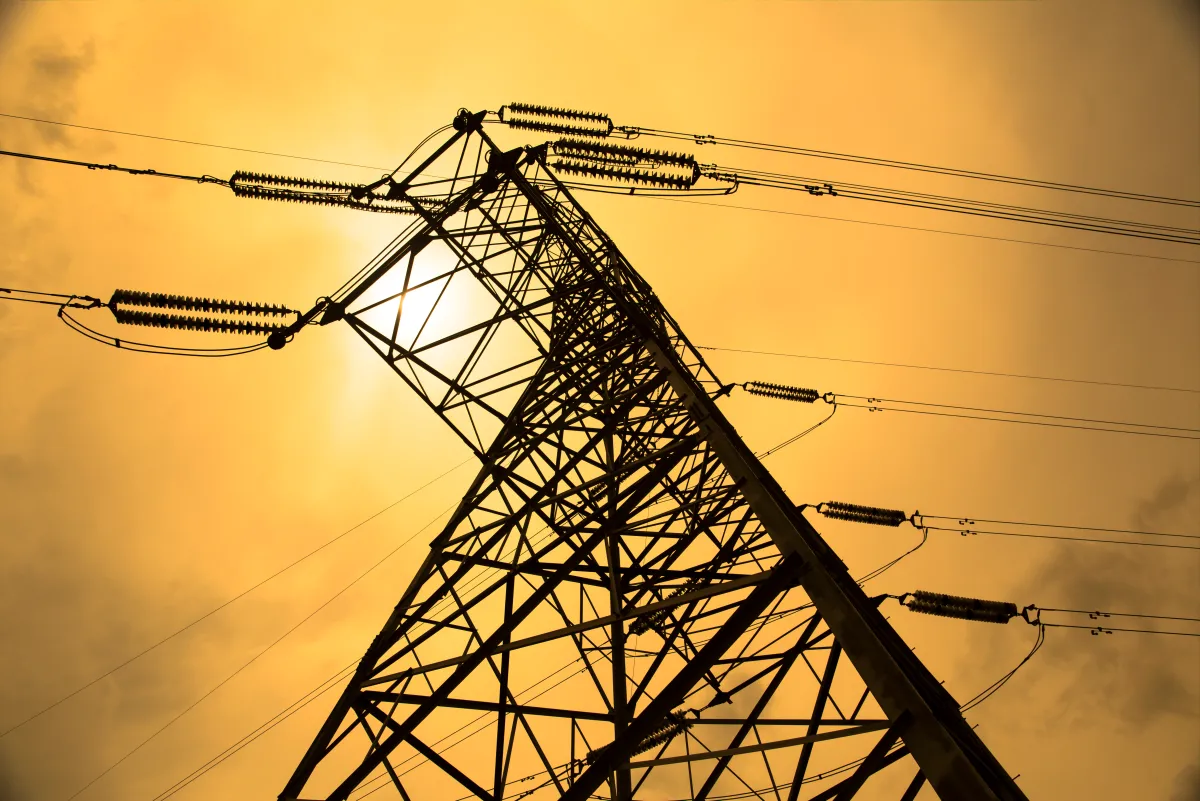 Electric transmission tower under a golden sky with visible power lines and insulators, sun partially obscured.