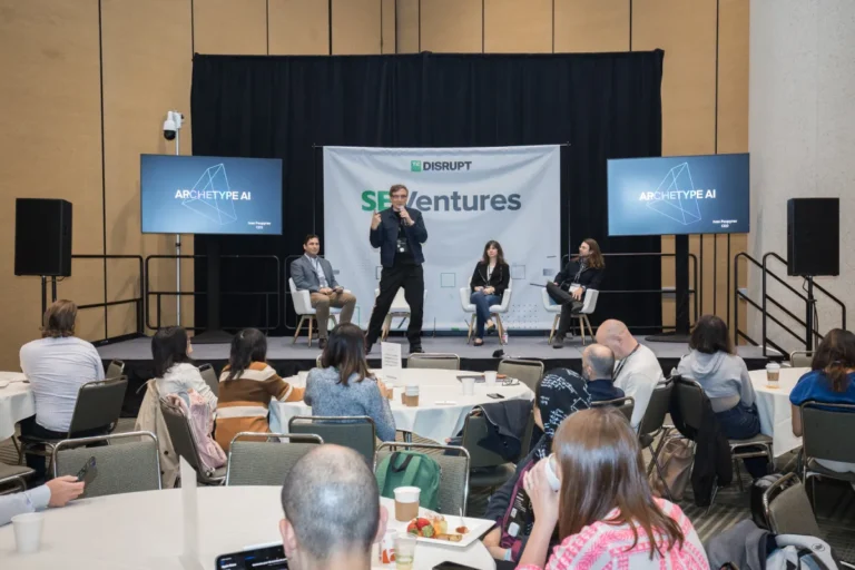 Person speaking on stage at a conference, three panelists seated, screens displaying "Archetype AI," audience in foreground.