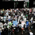 Crowded tech conference with numerous booths, people networking, and banners displaying company logos and startup informat...