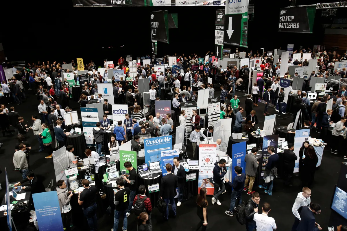 Crowded tech conference with numerous booths, people networking, and banners displaying company logos and startup informat...