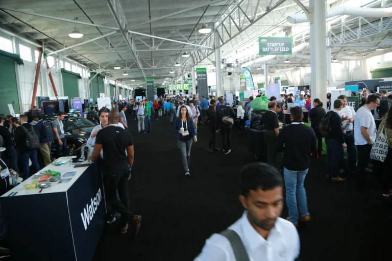 Crowded expo hall with booths and attendees, banners for "Startup Battlefield" visible, people engaged in conversations.