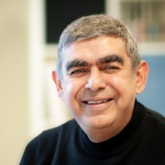 Smiling man with short gray hair and a black shirt in a blurred office setting.