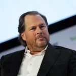 Man in a suit sits on a stage, looking thoughtful, with a blurred background displaying partial logos.