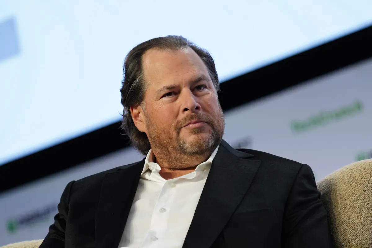 Man in a suit sits on a stage, looking thoughtful, with a blurred background displaying partial logos.