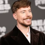 Smiling man in a dark suit at an event, with blurred "prime" and "beast games" logos in the background.
