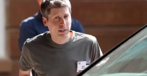 A man in a gray shirt with a name tag stands outdoors, partially obscured by a car, another person in the background.
