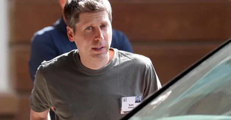 A man in a gray shirt with a name tag stands outdoors, partially obscured by a car, another person in the background.