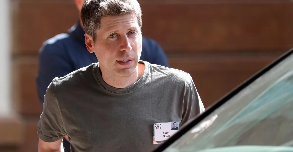 A man in a gray shirt with a name tag stands outdoors, partially obscured by a car, another person in the background.