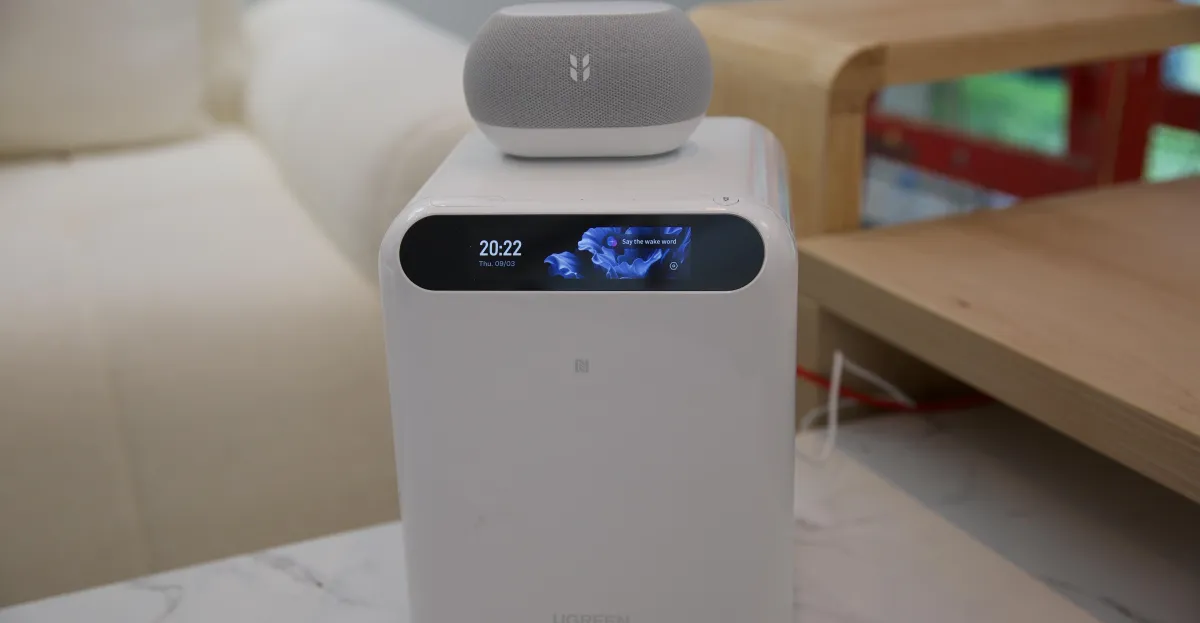 White smart home device with digital display showing time and date, small speaker on top, set on a marble table.