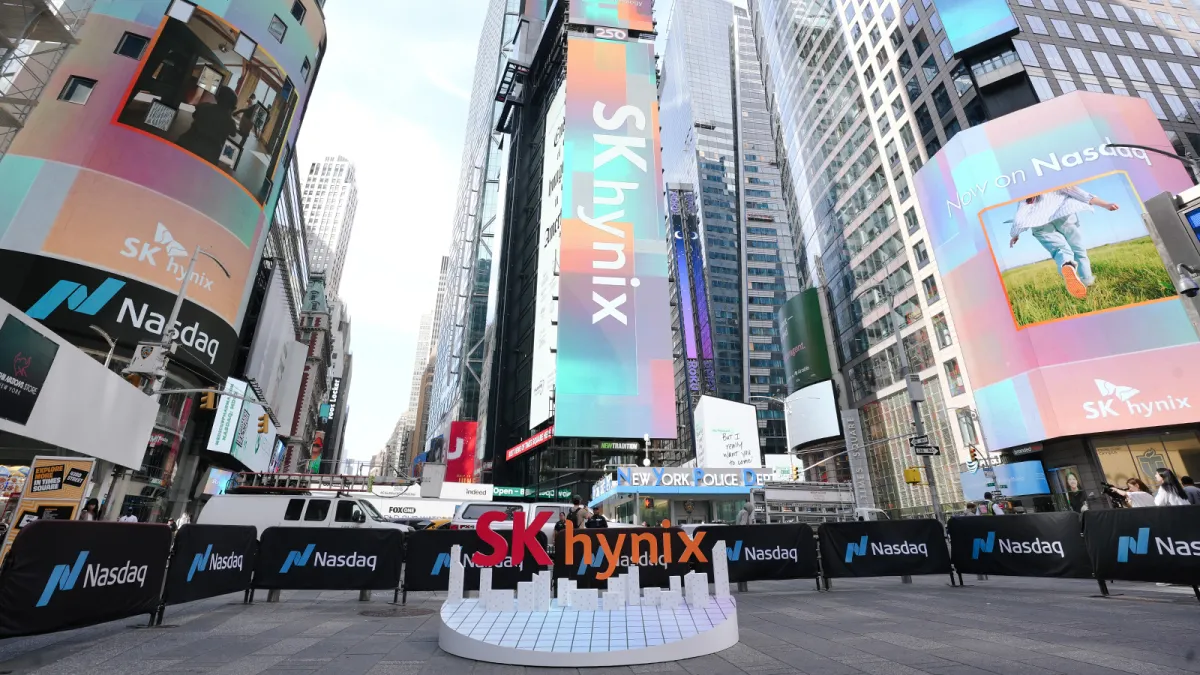 SK Hynix and Nasdaq logos displayed on digital billboards in a cityscape with skyscrapers and a small stage setup.