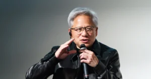 Person in a black leather jacket speaks into a microphone, gesturing with one hand against a plain background.