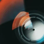 Close-up of a camera lens with orange and blue reflections, showing aperture blades in a circular pattern.