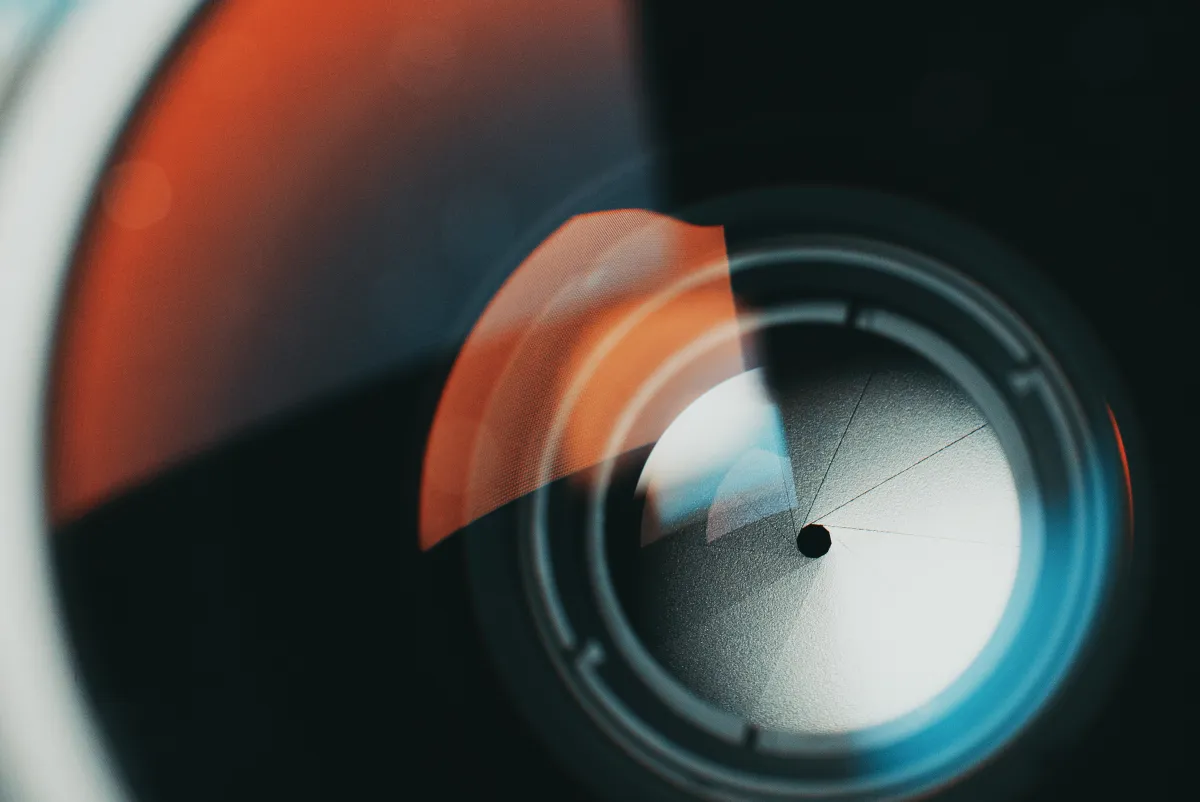 Close-up of a camera lens with orange and blue reflections, showing aperture blades in a circular pattern.