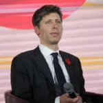 Man in a suit holding a microphone, seated on stage with a red and pink abstract background.
