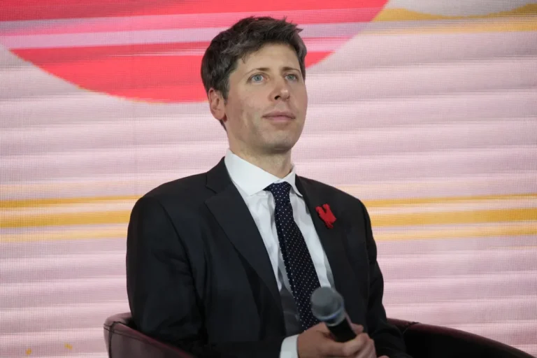 Man in a suit holding a microphone, seated on stage with a red and pink abstract background.