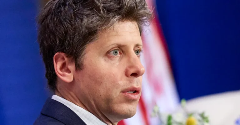 Person in a suit speaking, with an American flag in the blurred background.
