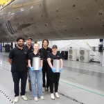 Five people in a hangar stand by a large rocket component, two holding cylindrical objects, wearing black shirts and gloves.