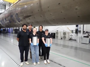 Five people in a hangar stand by a large rocket component, two holding cylindrical objects, wearing black shirts and gloves.