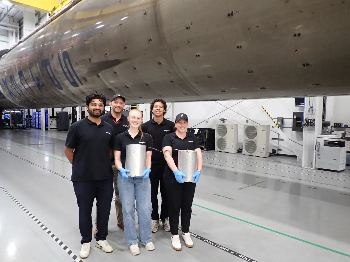 Five people in a hangar stand by a large rocket component, two holding cylindrical objects, wearing black shirts and gloves.