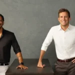 Two men smiling, one in a black shirt and the other in white, standing next to a dark table against a gray background.