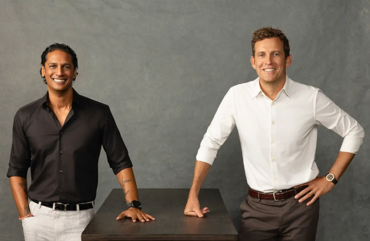 Two men smiling, one in a black shirt and the other in white, standing next to a dark table against a gray background.