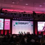 Conference stage with speakers and audience, large screens displaying "Disrupt" and "AI Stage powered by Google Cloud".