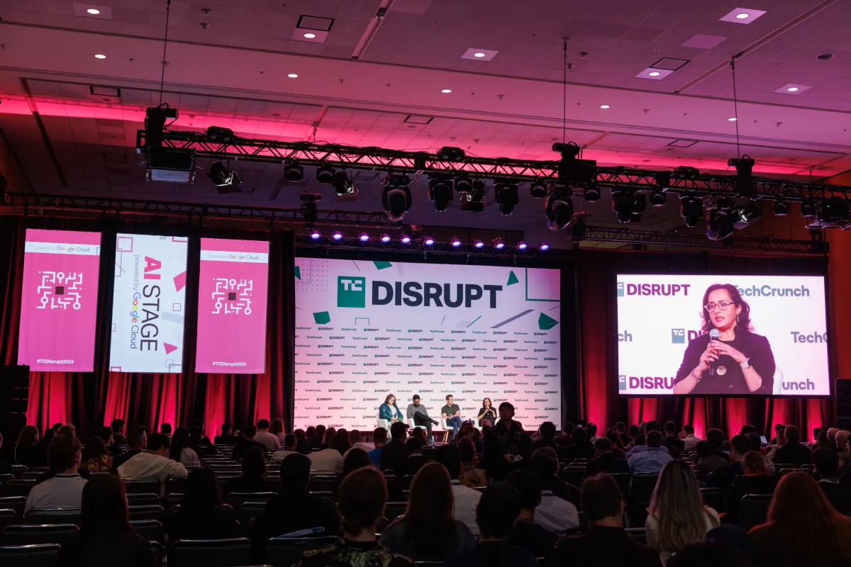 Conference stage with speakers and audience, large screens displaying "Disrupt" and "AI Stage powered by Google Cloud".