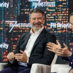 Three men in suits engaged in a lively discussion on stage with "FII Priority" displayed in the background.