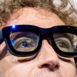 Close-up of a person wearing thick black smart glasses, reflecting light, with curly hair visible.