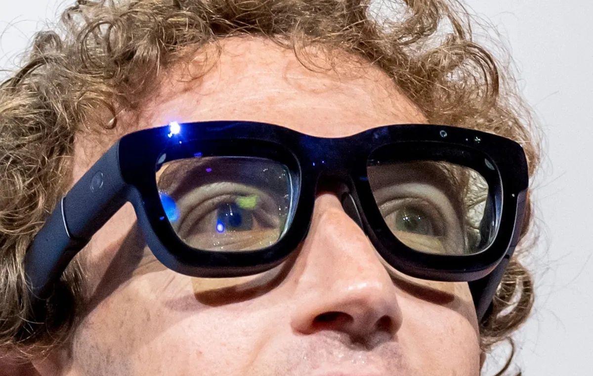 Close-up of a person wearing thick black smart glasses, reflecting light, with curly hair visible.