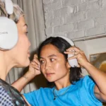 Two people wearing Sonos headphones in a living room, one adjusting their headphones, white brick wall in the background.