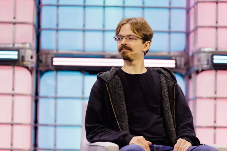 A person with glasses and a microphone sits on stage against a colorful geometric background.