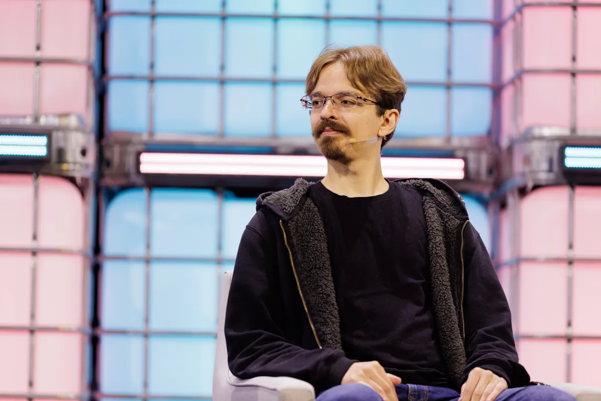 A person with glasses and a microphone sits on stage against a colorful geometric background.